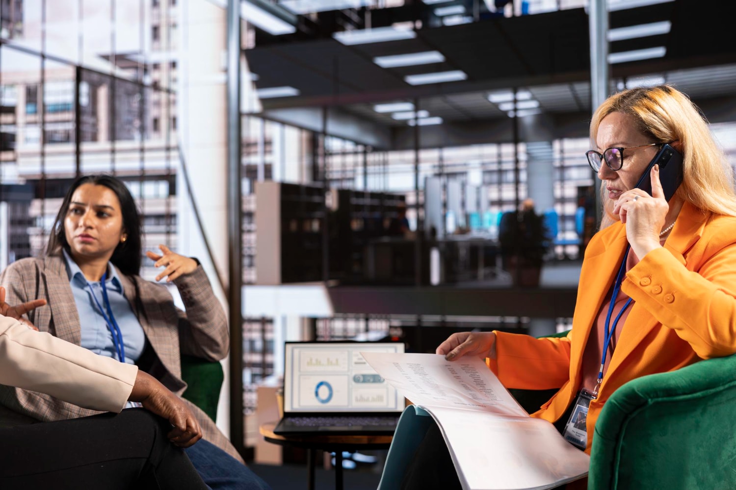 Three women have a dynamic conversation in a corporate lounge. One is on a mobile phone while reviewing printed documents. This illustrates the flexibility of business phone systems and how cloud phone systems support mobile VoIP phone solution access.