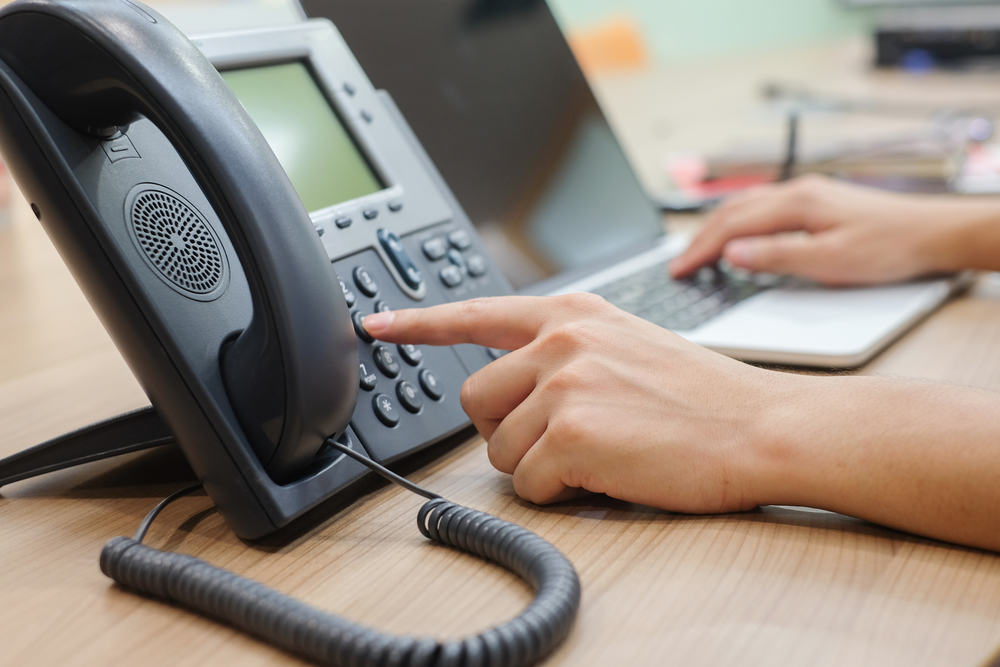 Close-up of a person dialing a desk phone while working on a laptop in an office setting