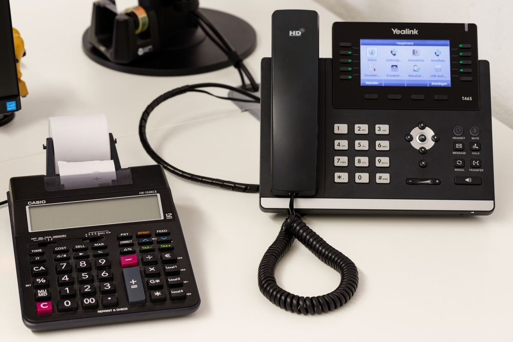 Yealink VoIP desk phone with HD display next to a printing calculator on office desk